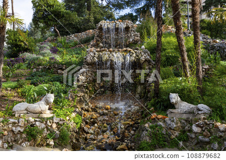 Waterfall. Waterfall in a park in Madrid. Artificial Mountain of Buen Retiro, called Mountain of the Cats. Dark stone background surrounded by green vegetation and plants and leaves. Autumn colors. Waterfall. Waterfall in a park in Madrid. Artificial Mountain of Buen Retiro, called Mountain of the Cats. Dark stone background surrounded by green vegetation and plants and leaves. Autumn colors. 108879682