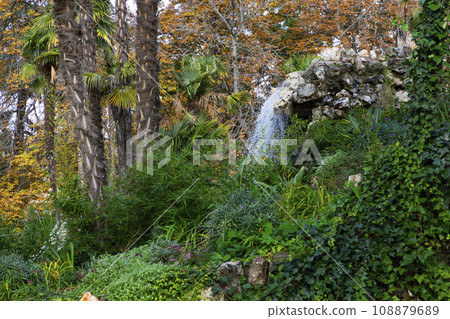 Waterfall. Waterfall in a park in Madrid. Artificial Mountain of Buen Retiro, called Mountain of the Cats. Dark stone background surrounded by green vegetation and plants and leaves. Autumn colors. 108879689