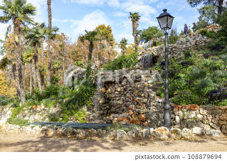 Waterfall. Waterfall in a park in Madrid. Artificial Mountain of Buen Retiro, called Mountain of the Cats. Dark stone background surrounded by green vegetation and plants and leaves. Autumn colors. 108879694