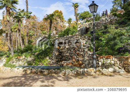 Waterfall. Waterfall in a park in Madrid. Artificial Mountain of Buen Retiro, called Mountain of the Cats. Dark stone background surrounded by green vegetation and plants and leaves. Autumn colors. 108879695