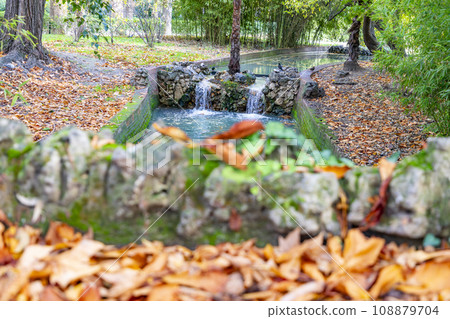 Waterfall. Waterfall in a park in Madrid. Artificial Mountain of Buen Retiro, called Mountain of the Cats. Dark stone background surrounded by green vegetation and plants and leaves. Autumn colors. 108879704