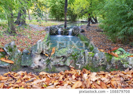 Waterfall. Waterfall in a park in Madrid. Artificial Mountain of Buen Retiro, called Mountain of the Cats. Dark stone background surrounded by green vegetation and plants and leaves. Autumn colors. 108879708