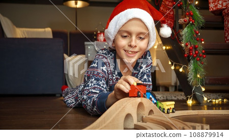 Portrait of happy little boy celebrating Christmas lying on floor and playing with colorful wooden train and wooden railroad. Winter holidays, celebrations and party. Portrait of happy little boy celebrating Christmas lying on floor and playing with colorful wooden train and wooden railroad. Winter holidays, celebrations and party. 108879914