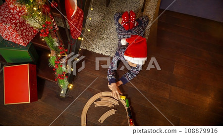 Cute boy in pajamas lying on floor and playing with toy wooden train and railroad while celebrating Christmas or New Year. Winter holidays, celebrations and party. 108879916