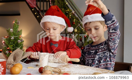 Two boys in Santa's hats celebrating Christmas making dough and playing with flour on kitchen. Winter holidays, celebrations and party. Two boys in Santa's hats celebrating Christmas making dough and playing with flour on kitchen. Winter holidays, celebrations and party. 108879963