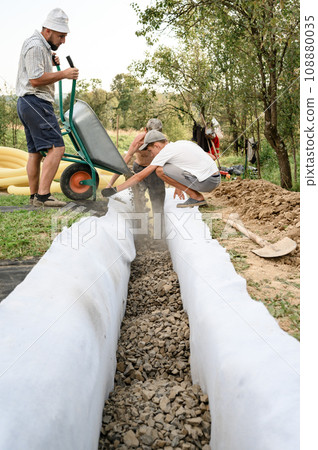 Man pours rubble from a wheelbarrow into a trench. Drainage works for drainage of ground water around the field. Man pours rubble from a wheelbarrow into a trench. Drainage works for drainage of ground water around the field. 108880035