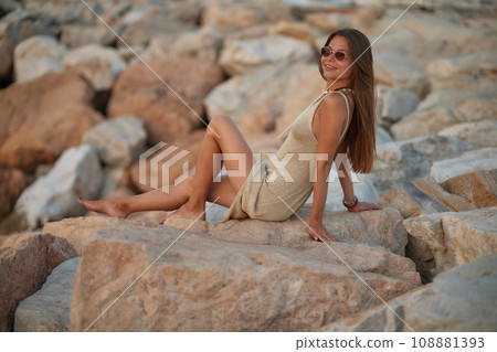Closeup portrait of attractive brunette girl with long hair posing on rocky beach on sunset background. She wears elegant long dress, sunglasses. She is smiling. Closeup portrait of attractive brunette girl with long hair posing on rocky beach on sunset background. She wears elegant long dress, sunglasses. She is smiling. 108881393