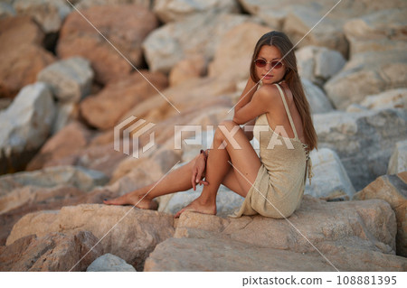 Closeup portrait of attractive brunette girl with long hair posing on rocky beach on sunset background. She wears elegant long dress, sunglasses. She is smiling. 108881395