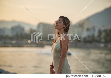 Cute stylish girl posing on the beach. The girl is dressed in a stylish off-the-shoulder dress. She's wearing brown sunglasses. The ocean is behind her. Cute stylish girl posing on the beach. The girl is dressed in a stylish off-the-shoulder dress. She's wearing brown sunglasses. The ocean is behind her. 108881820