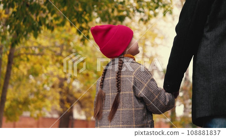 Little girl looks around walking with father in autumn garden. Curious little daughter holds father hand walking in park. Father and little daughter enjoy autumn day in yellow public urban garden Little girl looks around walking with father in autumn garden. Curious little daughter holds father hand walking in park. Father and little daughter enjoy autumn day in yellow public urban garden 108881837