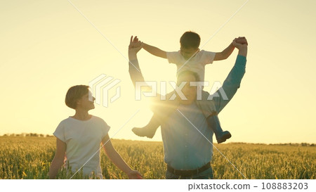 a little boy sits riding on his father's shoulders in a wheat field. dad farmer farming. happy family walks at sunset in the field. childhood dream happy family. child plays pilot flight sky. dream 108883203