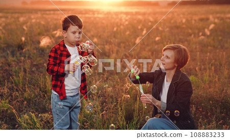 Boy with mother stands in field at sunset blowing soap bubbles into fiery sky Boy with mother stands in field at sunset blowing soap bubbles into fiery sky 108883233