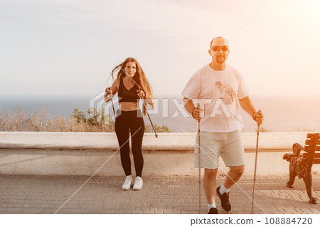 Happy Middle aged couple or friends practicing nordic walking in park near sea. Mature couple with trekking poles walking, practicing Nordic walking outdoors. Aging youthfully and sport concept 108884720