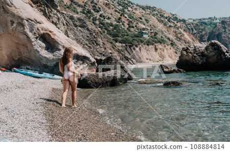Woman travel sea. Happy tourist in white bikini enjoy taking picture outdoors for memories. Woman traveler posing on the beach at sea surrounded by volcanic mountains, sharing travel adventure journey 108884814