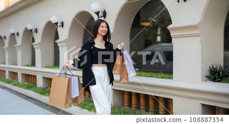 Happy beautiful young stylish woman asian with shopping bag while walking near mall on holiday Black Friday Happy beautiful young stylish woman asian with shopping bag while walking near mall on holiday Black Friday 108887354