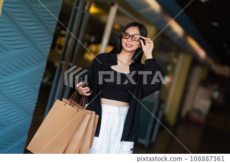 Woman using smartphone holding Black Friday shopping bag while standing on the side with the mall background 108887361