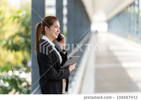 Smiling businesswoman with tablet talking on mobile phone going to work early in morning Smiling businesswoman with tablet talking on mobile phone going to work early in morning 108887432