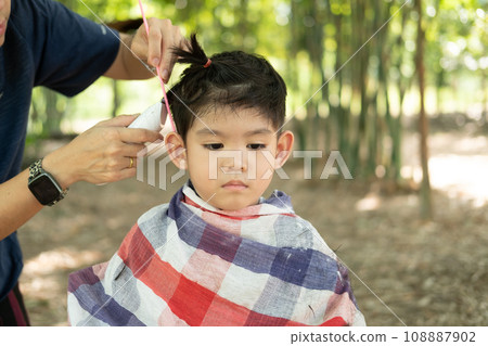 Barber cutting hair of an Asian boy In an open space filled with trees. 108887902