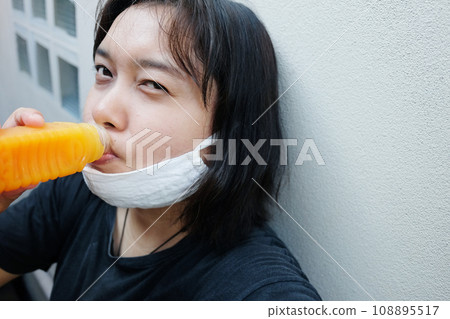 Asian Thai woman wearing a white cloth mask for prevent the Covid-19 or Corona virus and drinking orange juice in bottle for vitamin c helps to refresh. Air Pollution Value Pm 2.5 in Thailand. Asian Thai woman wearing a white cloth mask for prevent the Covid-19 or Corona virus and drinking orange juice in bottle for vitamin c helps to refresh. Air Pollution Value Pm 2.5 in Thailand. 108895517