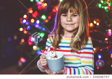 Little child girl holding cup with hot chocolate with marshmallows as Santa Claus. Kid by near Christmas tree decorated with lights. Little child girl holding cup with hot chocolate with marshmallows as Santa Claus. Kid by near Christmas tree decorated with lights. 108896747