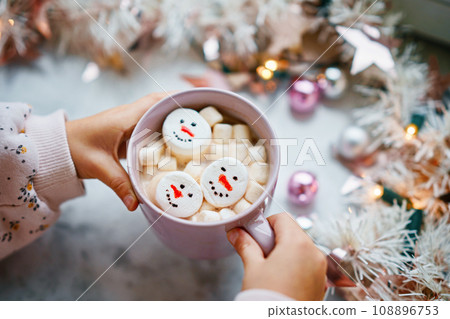 Unrecognizable little child girl holding cup with hot chocolate with marshmallows as snowman. Kid sitting near Christmas decorated window with lights. 108896753