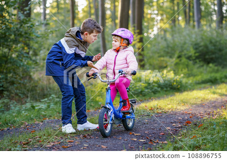 Cute little preschool girl in safety helmet riding bicycle. School kid boy, brother teaching happy healthy sister child cycling and having fun with learning bike. Active siblings family outdoors. 108896755
