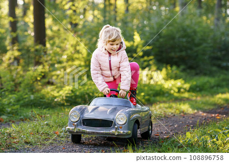 Little preschool girl driving big vintage toy car. Happy child having fun with playing outdoors. Active preschooler child enjoying warm autumn day in forest. Smiling stunning kid playing Little preschool girl driving big vintage toy car. Happy child having fun with playing outdoors. Active preschooler child enjoying warm autumn day in forest. Smiling stunning kid playing 108896758