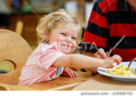 Toddler girl eating healthy vegetables and unhealthy french fries potatoes. Cute happy baby child taking food from parents dish in restaurant. Father eating in fast food restaurant with daughter Toddler girl eating healthy vegetables and unhealthy french fries potatoes. Cute happy baby child taking food from parents dish in restaurant. Father eating in fast food restaurant with daughter 108896792