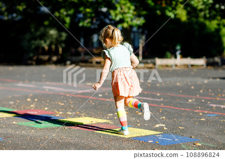 Cute little toddler girl playing hopscotch game drawn with colorful chalks on asphalt. Little active child jumping on playground outdoors on a sunny day. Summer activities for children. 108896824