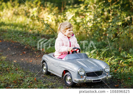 Little preschool girl driving big vintage toy car. Happy child having fun with playing outdoors. Active preschooler child enjoying warm autumn day in forest. Smiling stunning kid playing 108896825