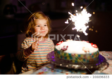 Adorable little toddler girl celebrating second birthday. Baby child eating marshmellows decoration on homemade cake, indoor. Happy healthy toddler is suprised about firework sparkler on cake Adorable little toddler girl celebrating second birthday. Baby child eating marshmellows decoration on homemade cake, indoor. Happy healthy toddler is suprised about firework sparkler on cake 108896833