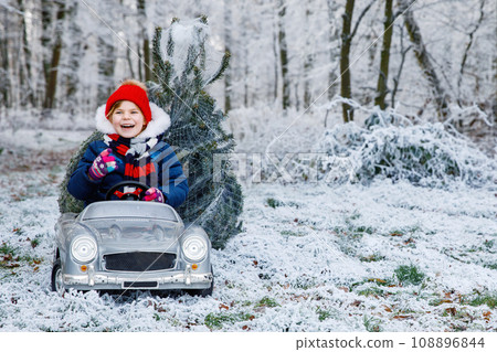 Happy little smiling girl driving toy car with Christmas tree. Funny preschool child in winter clothes bringing hewed xmas tree from snowy forest. Family, tradition, holiday. 108896844