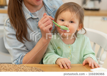 Happy family at home. Mother feeding her baby girl from spoon in kitchen. Little toddler child with messy funny face eats healthy food at home. Young woman mom giving food to kid daughter Happy family at home. Mother feeding her baby girl from spoon in kitchen. Little toddler child with messy funny face eats healthy food at home. Young woman mom giving food to kid daughter 108896966