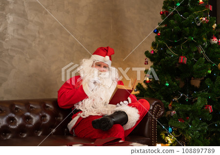 Santa claus reading red book while sitting on sofa in living room at home. Santa claus reading red book while sitting on sofa in living room at home. 108897789