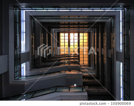 Stair in the square building from 1st floor shoots up to rooftop glass with the orange sunlight down on it. 108900069