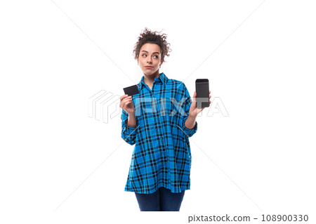 young smart curly brunette woman in blue plaid shirt shows plastic card and phone 108900330