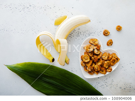 Dried banana slices in heart shape plate with raw banana and green leaf. 108904006