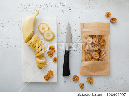 Pack of dried bio banana chip slices with knife and raw banana.Top view. 108904007