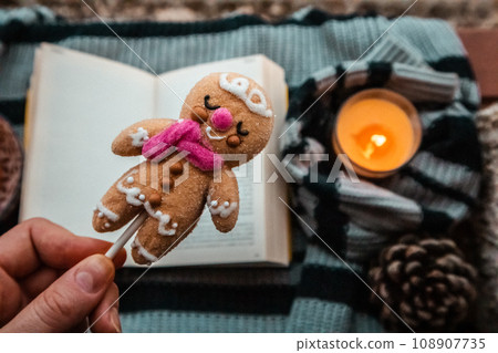 Homemade traditional Christmas cookies with icing in a hand. Delicious gingerbread man against open paper book. New Year 2024 doll, toy, sweet in a hand top view. Reading at home with orange candle. Homemade traditional Christmas cookies with icing in a hand. Delicious gingerbread man against open paper book. New Year 2024 doll, toy, sweet in a hand top view. Reading at home with orange candle. 108907735