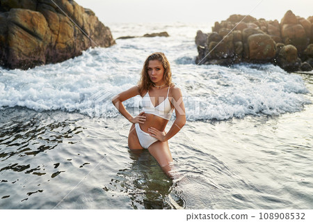 Beautiful woman poses in white bikini on seaside, rocks behind, wading in ocean water. Beautiful woman poses in white bikini on a seaside, rocks behind, wading in ocean water. Beautiful woman poses in white bikini on seaside, rocks behind, wading in ocean water. Beautiful woman poses in white bikini on a seaside, rocks behind, wading in ocean water. 108908532
