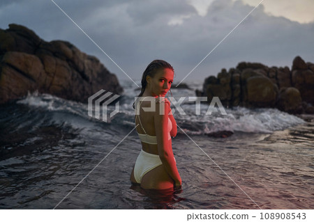 Attractive young woman enjoys nocturnal swim in ocean, illuminated by crimson light. Female beauty in stylish swimwear, night-time leisure at sea, tranquil waterscape surrounds. Attractive young woman enjoys nocturnal swim in ocean, illuminated by crimson light. Female beauty in stylish swimwear, night-time leisure at sea, tranquil waterscape surrounds. 108908543