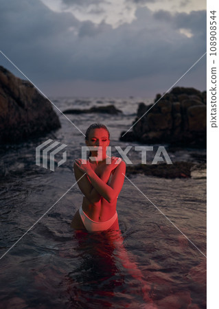 Attractive woman enjoys night swim in crimson lit ocean. Fashionable female basking in red illumination, sea backdrop, tranquil evening setting. Attractive woman enjoys night swim in crimson lit ocean. Fashionable female basking in red illumination, sea backdrop, tranquil evening setting. 108908544