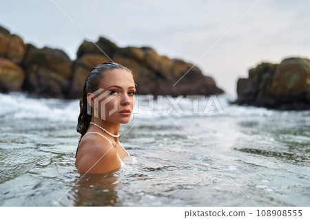 Woman with waterproof makeup submerges in ocean, stones backdrop. Female tours seascape, enduring swimwear stylish jewelry. Adventurous traveler explores coastal region, water resistant cosmetics. Woman with waterproof makeup submerges in ocean, stones backdrop. Female tours seascape, enduring swimwear stylish jewelry. Adventurous traveler explores coastal region, water resistant cosmetics. 108908555