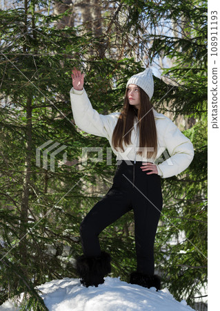 Teenager girl stands in winter forest, one raised hand holds pine branch, other hand placed on waist Teenager girl stands in winter forest, one raised hand holds pine branch, other hand placed on waist 108911193