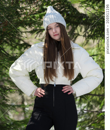 Portrait teenager girl arms akimbo, looking at camera. Fashion model standing in winter pine forest Portrait teenager girl arms akimbo, looking at camera. Fashion model standing in winter pine forest 108911196