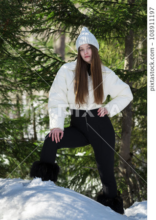 Teenager girl standing in winter forest in snowdrift on background of pine trees, looking at camera Teenager girl standing in winter forest in snowdrift on background of pine trees, looking at camera 108911197
