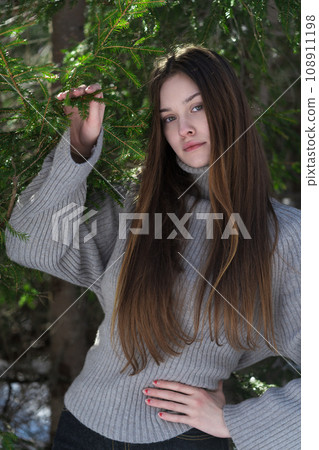 Teenager girl looking at camera, holding hand on waist, other raised and holds branch in pine forest Teenager girl looking at camera, holding hand on waist, other raised and holds branch in pine forest 108911198
