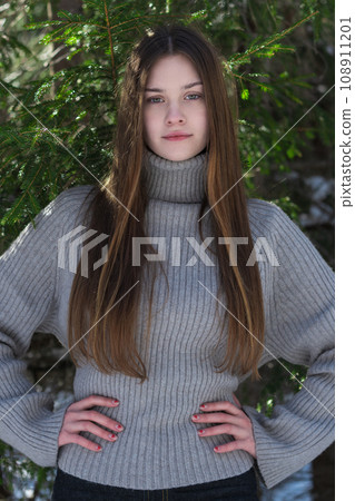 Teenage girl arms akimbo and looking at camera. Teen wearing in sweater standing in pine forest Teenage girl arms akimbo and looking at camera. Teen wearing in sweater standing in pine forest 108911201