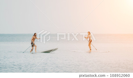Sea woman and man on sup. Silhouette of happy young woman and man, surfing on SUP board, confident paddling through water surface. Idyllic sunset. Active lifestyle at sea or river. 108912060
