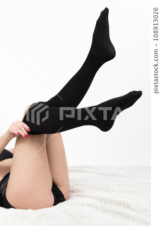 Female lies on bed, putting black pantyhose on slim legs, pulled up. Crop view unrecognizable woman 108913026